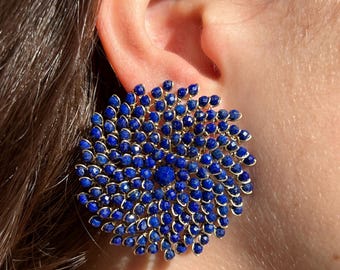 May include: A close-up of a large, round earring with a gold-colored frame. The earring is covered in small, deep blue gemstones, arranged in a radial pattern. The earring is worn on an ear.