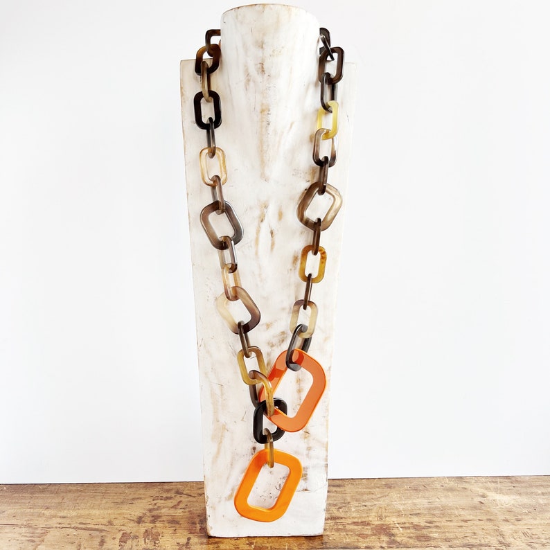 May include: A long necklace made of brown, black, and yellow rectangular links. The necklace is hanging on a white mannequin. The necklace has two large orange rectangular links hanging from the bottom.