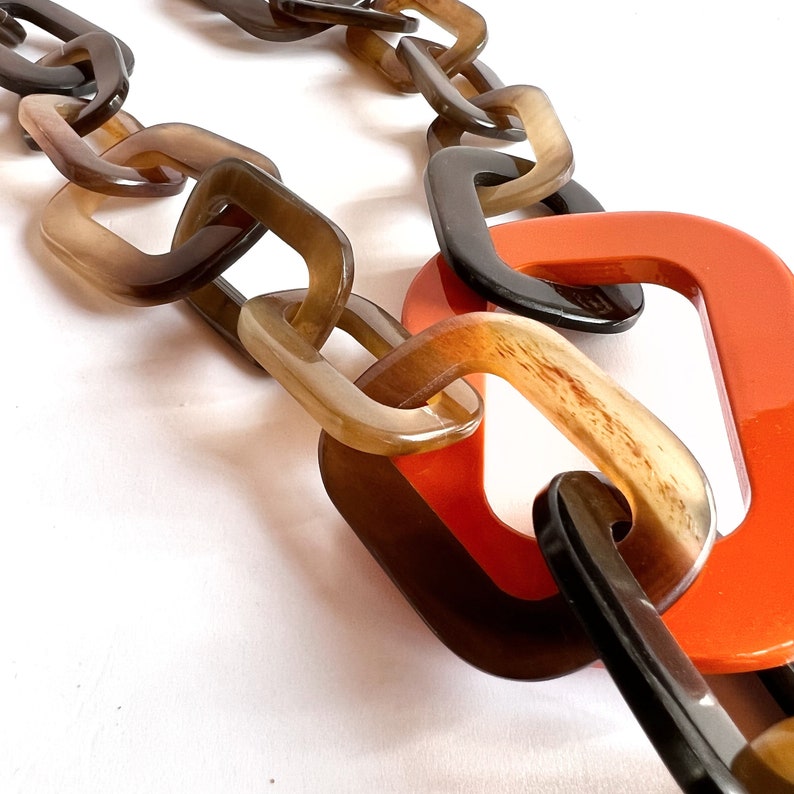 May include: A close-up of a chunky chain necklace made of brown and black plastic links. The chain is linked together with a bright orange plastic link.
