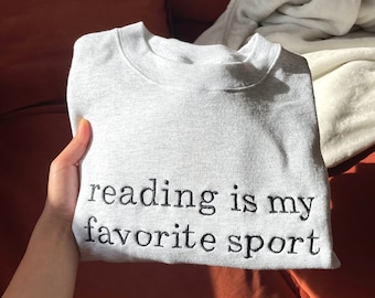 Könnte beinhalten: Hellgraues Sweatshirt mit dem in Schwarz gestickten Text "reading is my favorite sport". Das gefaltete Sweatshirt wird in einer Hand gehalten, mit einem rot-orangen Sofa im Hintergrund.