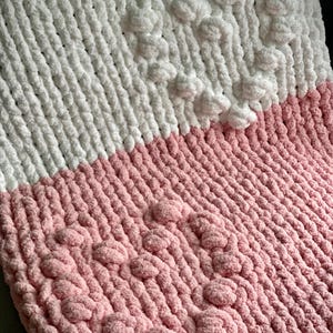 May include: A handmade, two-tone blanket in white and pink. The top half is white, and the bottom half is pink. Both sections have a textured, chunky knit design. The blanket features raised, textured, heart-shaped patterns in both colors.