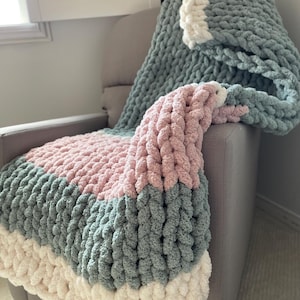 Peut inclure: Une couverture en tricot épais avec un motif rayé dans les tons de rose, gris et blanc. La couverture est drapé sur un fauteuil gris.