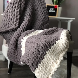 Può includere: Una coperta a maglia grossa grigia e bianca drappeggiata su una sedia. La coperta presenta una trama spessa e strutturata con un design a righe. Le parole "LIVE INSPIRED" sono su una piccola lavagna sullo sfondo.