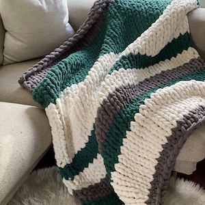 Può includere: Una coperta in maglia grossa con strisce di filato bianco, grigio scuro e verde bosco. La coperta è drappeggiata su un divano.