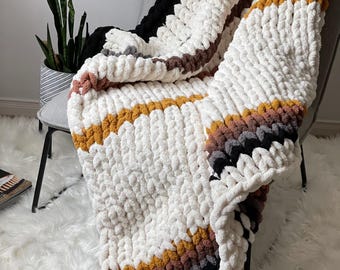 May include: A chunky knit throw blanket with stripes of white, black, brown, and mustard yellow. The blanket is draped over a chair.