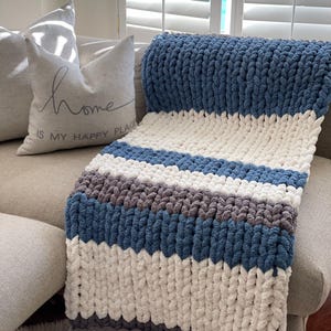 Op de afbeelding: Een dik gebreid deken met strepen van wit, grijs en blauw garen. Het deken is gedrapeerd over een bank met een kussen waarop "Home is my happy place" staat.