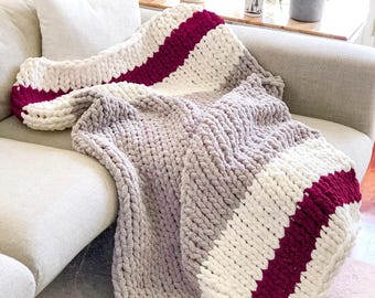 May include: A chunky knit throw blanket featuring horizontal stripes in cream, light gray, and deep magenta. The blanket is crafted from thick, soft yarn with a visible looped texture, draped over a neutral-colored sofa with a white throw pillow.