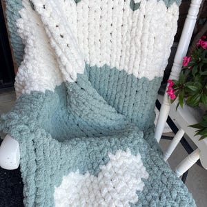 Puede incluir: Una manta de punto grueso en blanco y verde agua, sobre una mecedora blanca. La manta presenta un diseño de corazón y rayas horizontales. La manta suave y texturizada es un elemento de decoración acogedor.