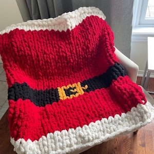 Könnte beinhalten: Eine rot-weiß-schwarze gestrickte Decke mit einer gelben Gürtelschnalle. Die Decke ist aus dickem Garn gefertigt und hat ein Weihnachtsmann-Motiv.