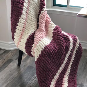 Puede incluir: Una manta de punto grueso con rayas de color burdeos, blanco y rosa. La manta está drapeada sobre una silla.