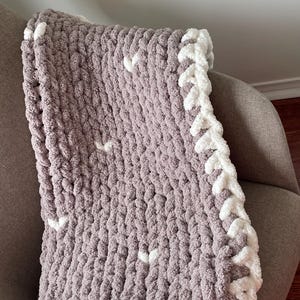 May include: A handmade, chunky knit blanket in a soft grey colour with white accents. The blanket features a white border and scattered white details. This cosy blanket is perfect for snuggling up or adding warmth to any room.