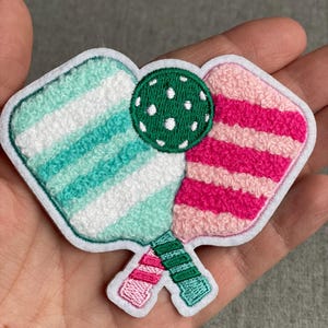 May include: An embroidered patch featuring two crossed pickleball paddles and a green pickleball with white dots. One paddle has light blue and white stripes, while the other has hot pink and light pink stripes. The patch has a white border.