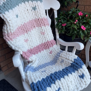 Könnte beinhalten: Eine grob gestrickte Decke in Weiß, Blau, Rosa und Grau liegt über einem weißen Schaukelstuhl. Die Decke hat ein gestreiftes Muster und ein weiches, strukturiertes Aussehen. Der Stuhl steht auf einer Veranda mit einer Backsteinmauer und Topfblumen.
