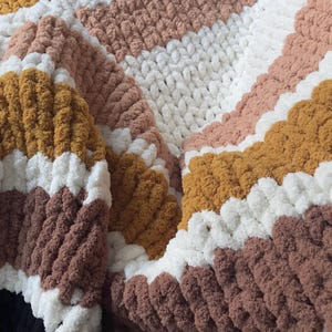 May include: A cozy, handmade blanket with a chunky knit design. The blanket features alternating stripes of white, brown, mustard yellow, and blush pink. The soft, textured material is perfect for snuggling up on the couch.