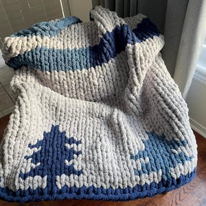 Puede incluir: Una manta de punto grueso en tonos grises y azules. La manta presenta un patrón a rayas con un gran diseño azul en el centro.