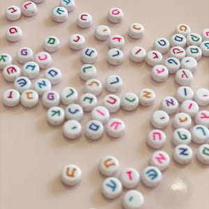 May include: A collection of small, round, white plastic beads, each featuring a single Hebrew letter printed in various colours including blue, red, green, orange, and purple. The beads are scattered across the frame, showing different characters.