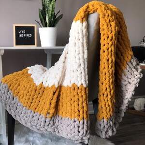 May include: A chunky knit throw blanket with stripes of mustard yellow, white and grey. The blanket is draped over a chair with a sign that reads "Live Inspired".