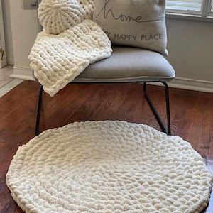 May include: A cream-colored, chunky knit rug sits on a hardwood floor. A matching throw blanket, pillow, and decorative ball are on a gray chair. The pillow has the word "home" and "my happy place" printed on it.