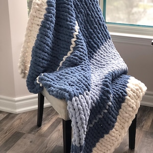 May include: A chunky knit blanket in shades of blue and white is draped over a chair. The blanket features a textured, oversized stitch pattern. The color scheme transitions from a deep blue to a lighter blue and white, creating a cozy and inviting aesthetic.