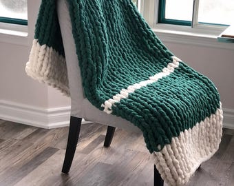May include: A chunky knit throw blanket in a deep green colour with white stripes. The blanket is draped over a chair.