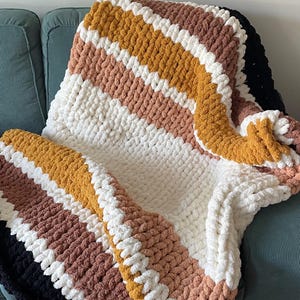 Peut inclure: Un plaid en tricot épais composé de fils doux et volumineux, présentant des rayures horizontales crème, jaune moutarde, rose poudré et marron, avec une section noire unie sur le bord. La couverture a un aspect texturé et bouclé.