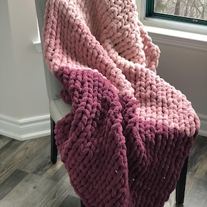 May include: A chunky knit throw blanket with a gradient of pink and burgundy colors. The blanket is draped over a chair.