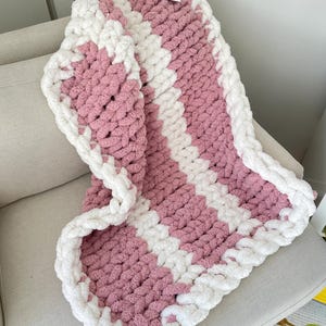 May include: A pink and white striped knitted baby blanket with a white border. The blanket is made with chunky yarn and has a soft, fluffy texture.