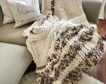 May include: A chunky knit throw blanket with a white base and brown and beige stripes. The blanket is draped over a sofa with a cushion that reads "Home is my happy place".