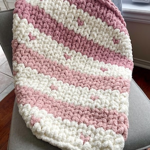 May include: A soft, chunky knit blanket with alternating stripes of white and pale pink. The blanket is draped over a grey chair, highlighting its texture and colour scheme. This cosy, handmade blanket is perfect for snuggling.