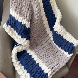 Puede incluir: Una manta de punto grueso en gris, blanco y azul marino, sobre un sillón gris. La manta presenta un hilo grueso y suave y un diseño a rayas. Esta acogedora manta es perfecta para acurrucarse en el sofá.