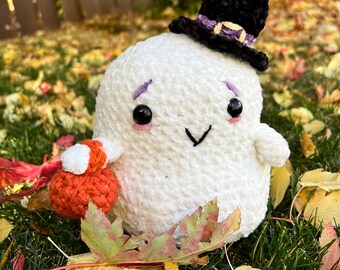 May include: A white crocheted ghost plushie featuring a black witch hat with a purple band and buckle. The ghost has black safety eyes, embroidered purple eyebrows, pink cheeks, and a black mouth. It holds a small orange crocheted pumpkin bag.