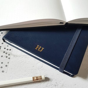 Könnte beinhalten: Ein marineblaues Notizbuch mit goldenen Initialen "HJ" und einem elastischen Verschluss. Im Hintergrund ist ein offenes Notizbuch mit gepunkteten Seiten sowie ein weißer Bleistift zu sehen.