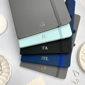 Peut inclure: Une pile de carnets personnalisés de différentes couleurs : gris, vert menthe, noir, bleu marine et gris. Chaque carnet présente des initiales en feuille métallique et une fermeture élastique. Les carnets sont empilés sur une surface blanche.