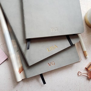 May include: Three grey notebooks with embossed initials: "BG", "LISA", and "VG". The "LISA" initials are gold. Each notebook has a black ribbon bookmark. A pencil, paper, and stationery are also visible.