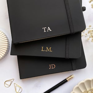 May include: A stack of three black notebooks with elastic closures. The top notebook has the initials "TA" in silver, the middle one has "L.M." in gold, and the bottom one has "JD" in gold. A pencil and gold paperclips are also visible.