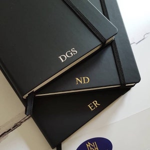 Black A5 notebooks with gold or silver personalised initials on the bottom of the journal cover. Each notebook has an elastic closure and pen loop.