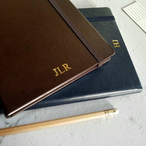 Könnte beinhalten: Zwei ledergebundene Notizbücher mit Gummibändern. Das obere Notizbuch ist braun mit den Initialen "JLR" in Gold geprägt. Das untere Notizbuch ist dunkelblau mit den Initialen "HJ" in Gold geprägt.