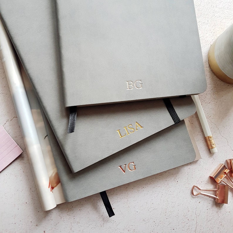 May include: A stack of three gray notebooks with personalized initials. The top notebook has "BG" embossed, the middle one reads "LISA" in gold, and the bottom one has "VG". Each notebook has a black ribbon bookmark.