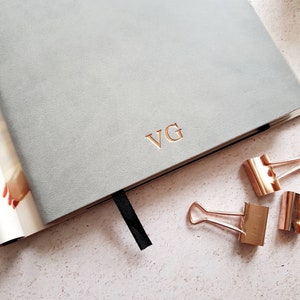 May include: A light gray journal with the initials "VG" in gold lettering. A black ribbon bookmark is visible. Three gold binder clips are scattered on the surface, suggesting an office or stationery theme.