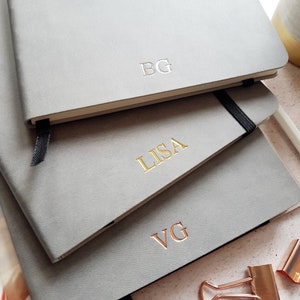 May include: Three gray notebooks stacked on top of each other. The top notebook has the initials "BG" embossed in the center. The middle notebook has the name "LISA" embossed in gold. The bottom notebook has the initials "VG" embossed in gold.
