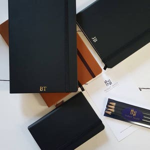 Black A5 notebooks with gold or silver personalised initials on the bottom of the journal cover. Each notebook has an elastic closure and pen loop.
