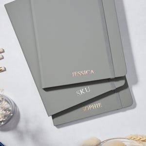 Può includere: Tre quaderni grigi impilati uno sopra l'altro. Il quaderno in alto ha il nome "JESSICA" scritto in lettere dorate. Il quaderno nel mezzo ha il nome "SKU" scritto in lettere argentate. Il quaderno in basso ha il nome "SOPHIE" scritto in lettere argentate.