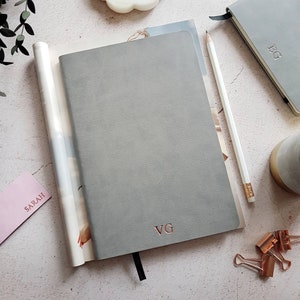 May include: A collection of gray notebooks and stationery. The main notebook has the initials "VG" in rose gold. A white pencil, a pink card with "SARAH" printed on it, and a small notebook with "FG" are also visible. The scene is set on a light-colored surface.