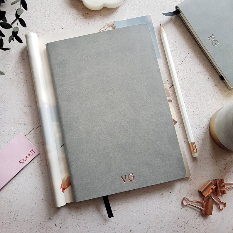 May include: A collection of gray notebooks and stationery. The main notebook has the initials "VG" in rose gold. A white pencil, a pink card with "SARAH" printed on it, and a small notebook with "FG" are also visible. The scene is set on a light-colored surface.