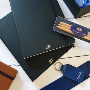 Black A5 notebooks with gold or silver personalised initials on the bottom of the journal cover. Each notebook has an elastic closure and pen loop.