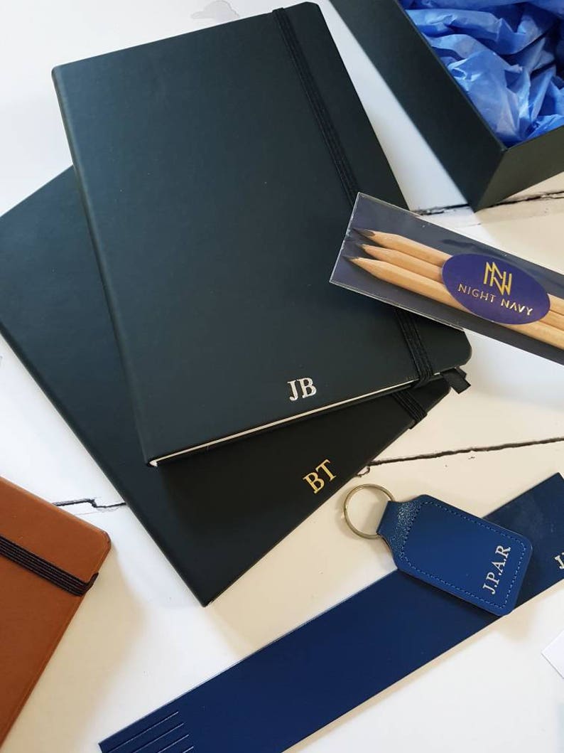 Black A5 notebooks with gold or silver personalised initials on the bottom of the journal cover. Each notebook has an elastic closure and pen loop.