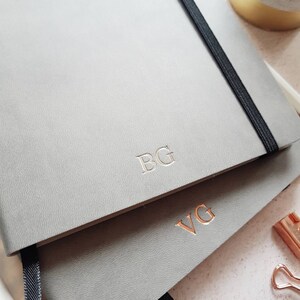 May include: Two gray notebooks with elastic closures are stacked. The top notebook has the initials "BG" embossed in silver, while the bottom notebook has "VG" in rose gold. The notebooks have a textured surface and a black elastic band.