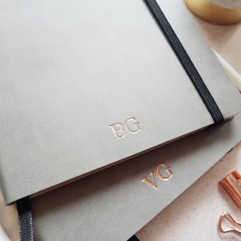 May include: Two gray notebooks with elastic closures are stacked. The top notebook has the initials "BG" embossed in silver, while the bottom notebook has "VG" in rose gold. The notebooks have a textured surface and a black elastic band.