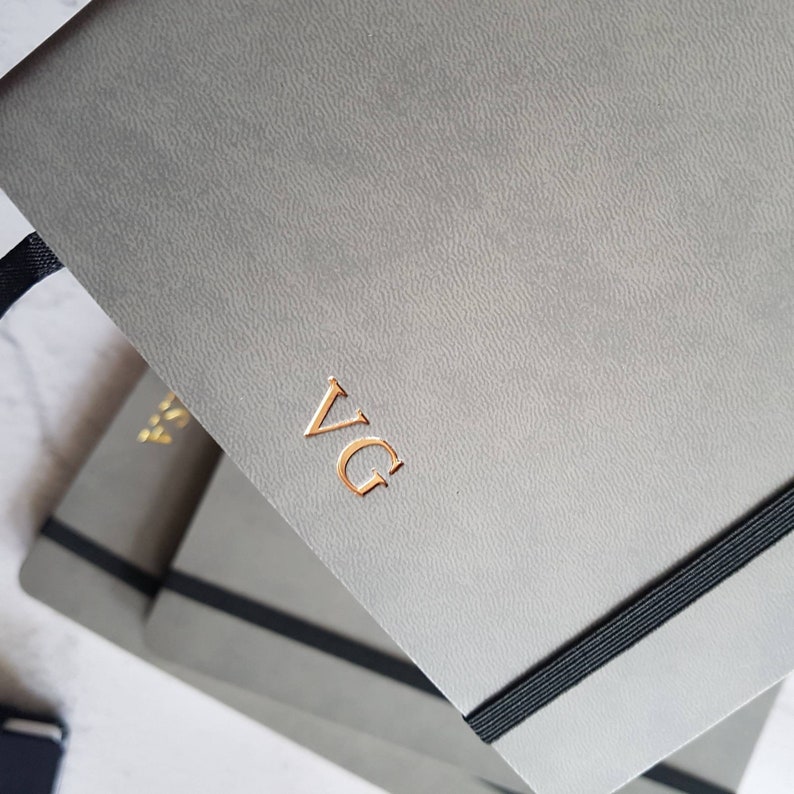 May include: Gray journal with gold-colored initials "VG" embossed on the cover. The journal has a textured surface, a black elastic band closure, and a black ribbon bookmark. The journal is part of a set.
