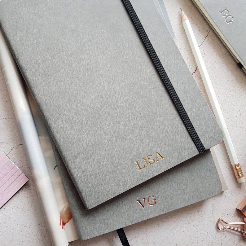 May include: Gray notebooks with gold and rose gold lettering. One notebook has "LISA" in gold, another has "VG" in rose gold. A white pencil and a black elastic band are visible.